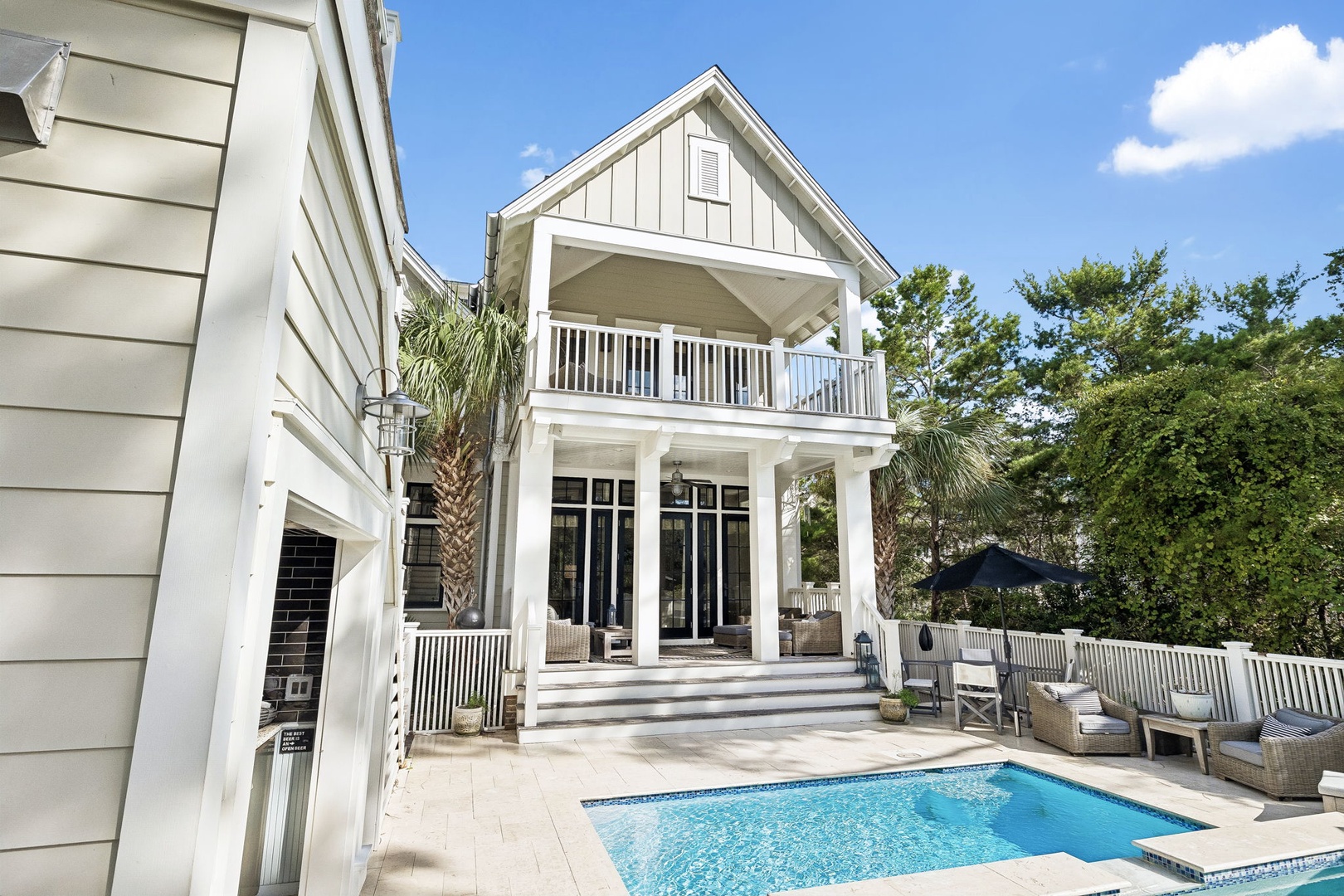 The pristine white facade overlooks a sparkling private pool framed by palms and lush greenery, with a spacious upper balcony offering an inviting retreat beneath sunny skies.
