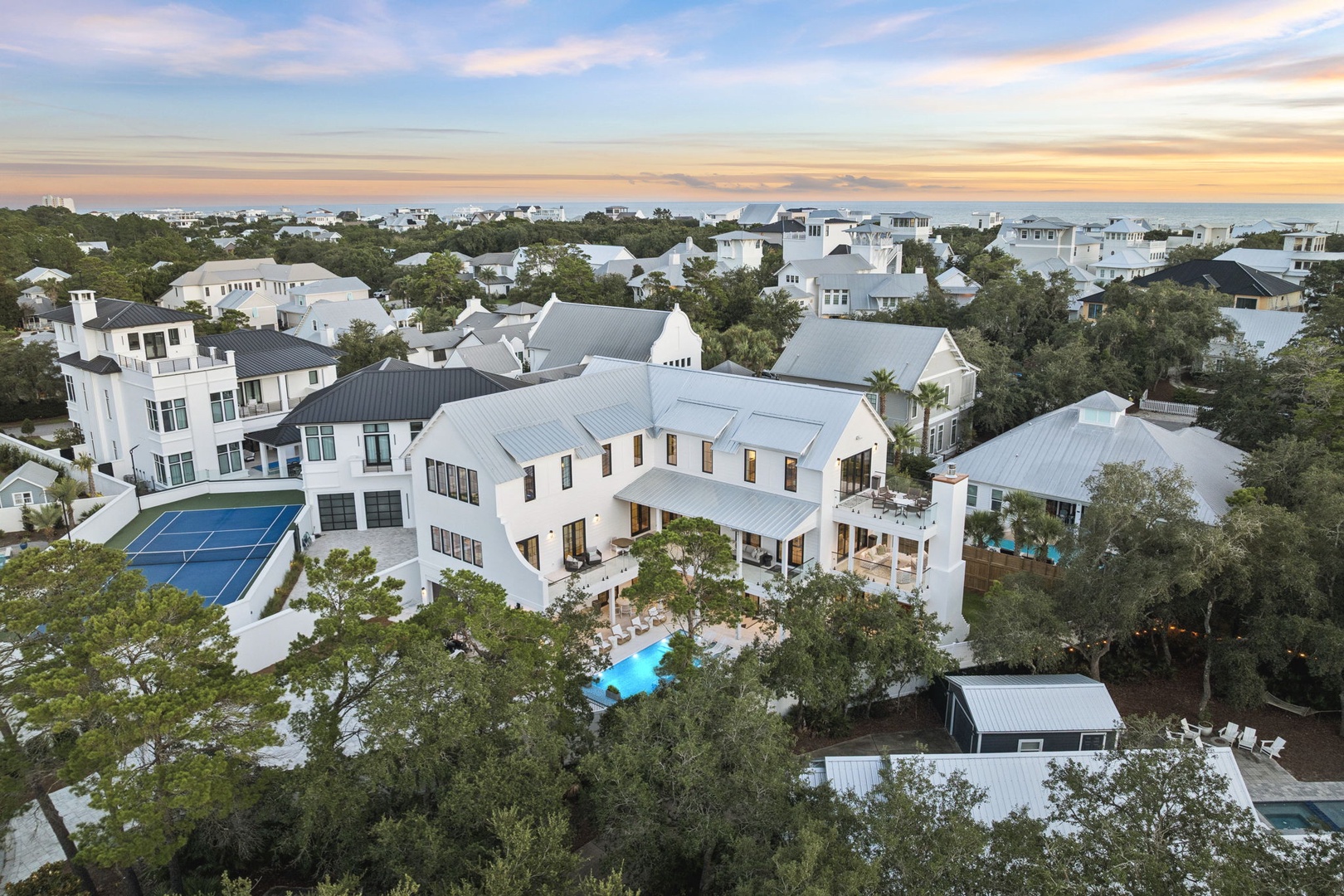 An expansive luxury estate nestled among coastal trees and neighboring homes, featuring a resort-style pool and generous outdoor living spaces beneath a painted evening sky.