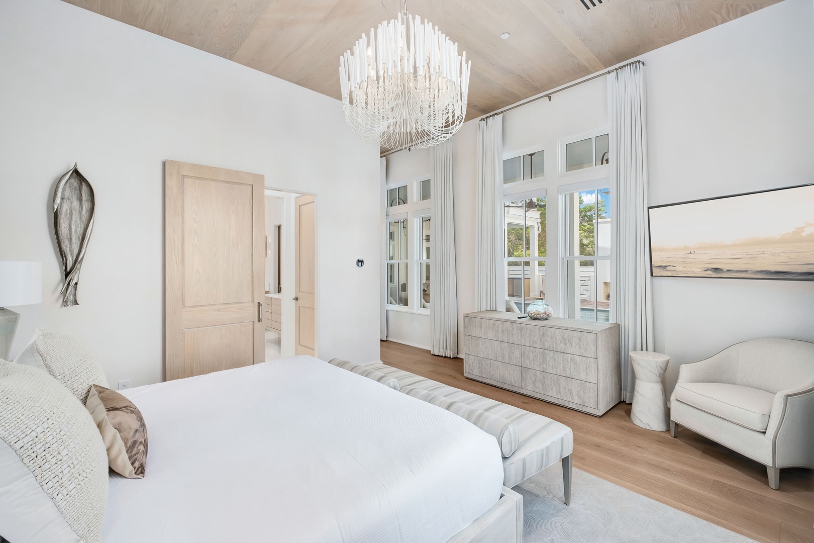 Step into your serene retreat where soaring ceilings and an elegant chandelier frame your plush bed. Natural light pours through tall windows, highlighting the soft textures and warm wood tones that make this space feel like a true sanctuary.
