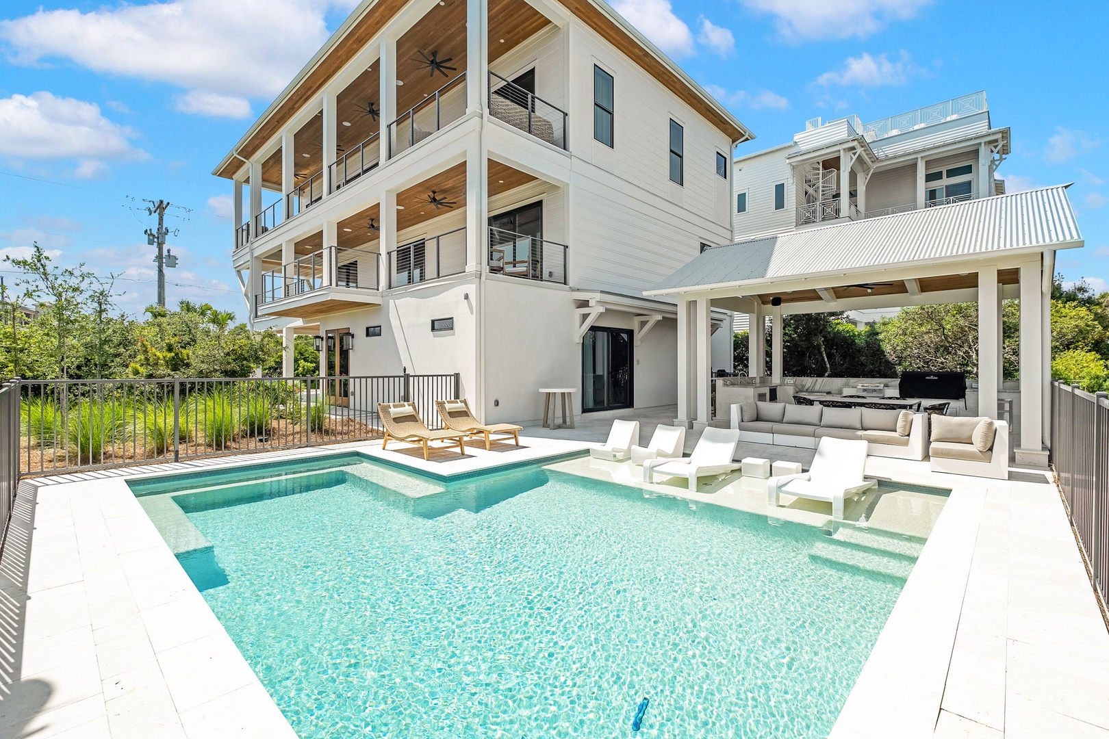 Modern coastal architecture featuring multiple covered decks, private pool with sun shelf, and outdoor lounge pavilion set among lush tropical landscaping under blue skies.