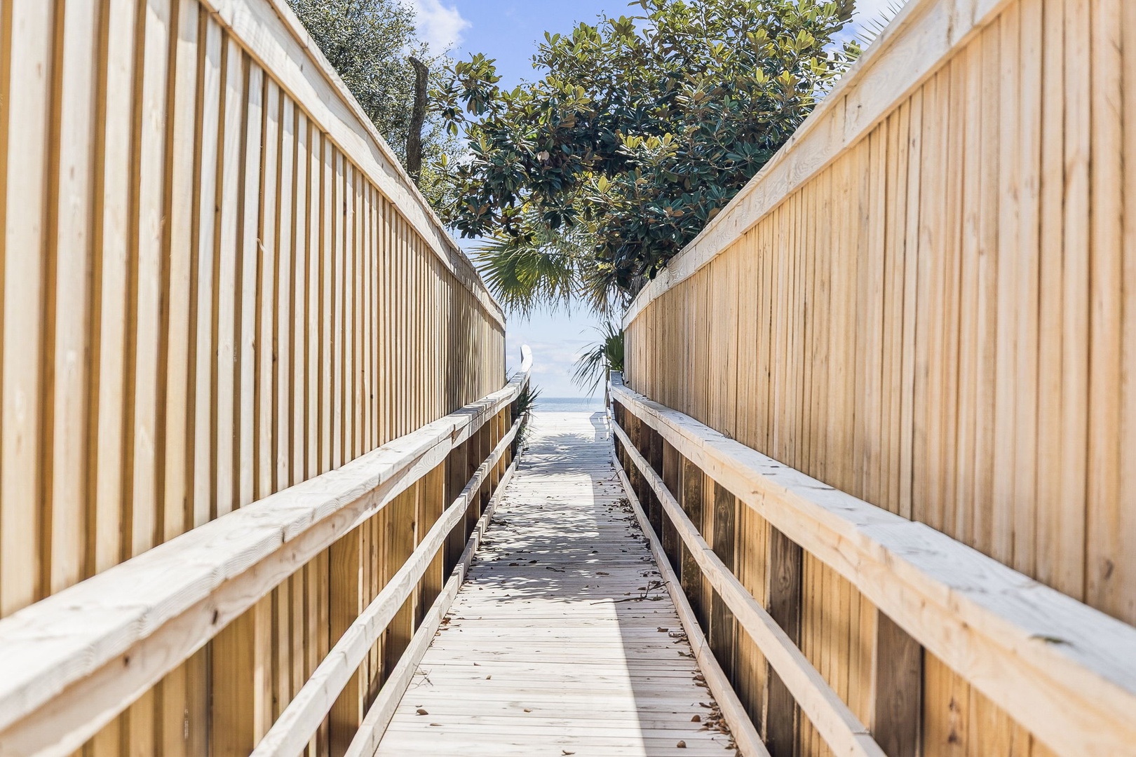 A wooden boardwalk leads through tropical vegetation to pristine beach access, framed by lush palms and azure skies.