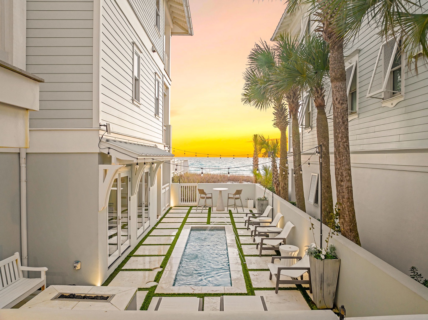 A serene courtyard pool framed by swaying palms and sunlit coastal views, steps from the sandy shore as day ends in golden hues.