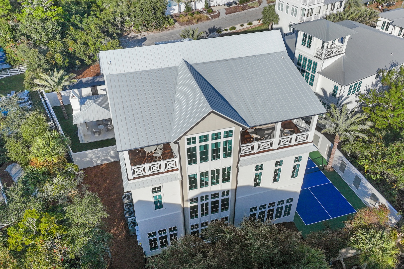 Elegant vacation rental with coastal architecture, multiple private balconies, and backyard amenities including a pool and sports court in a lush residential neighborhood.