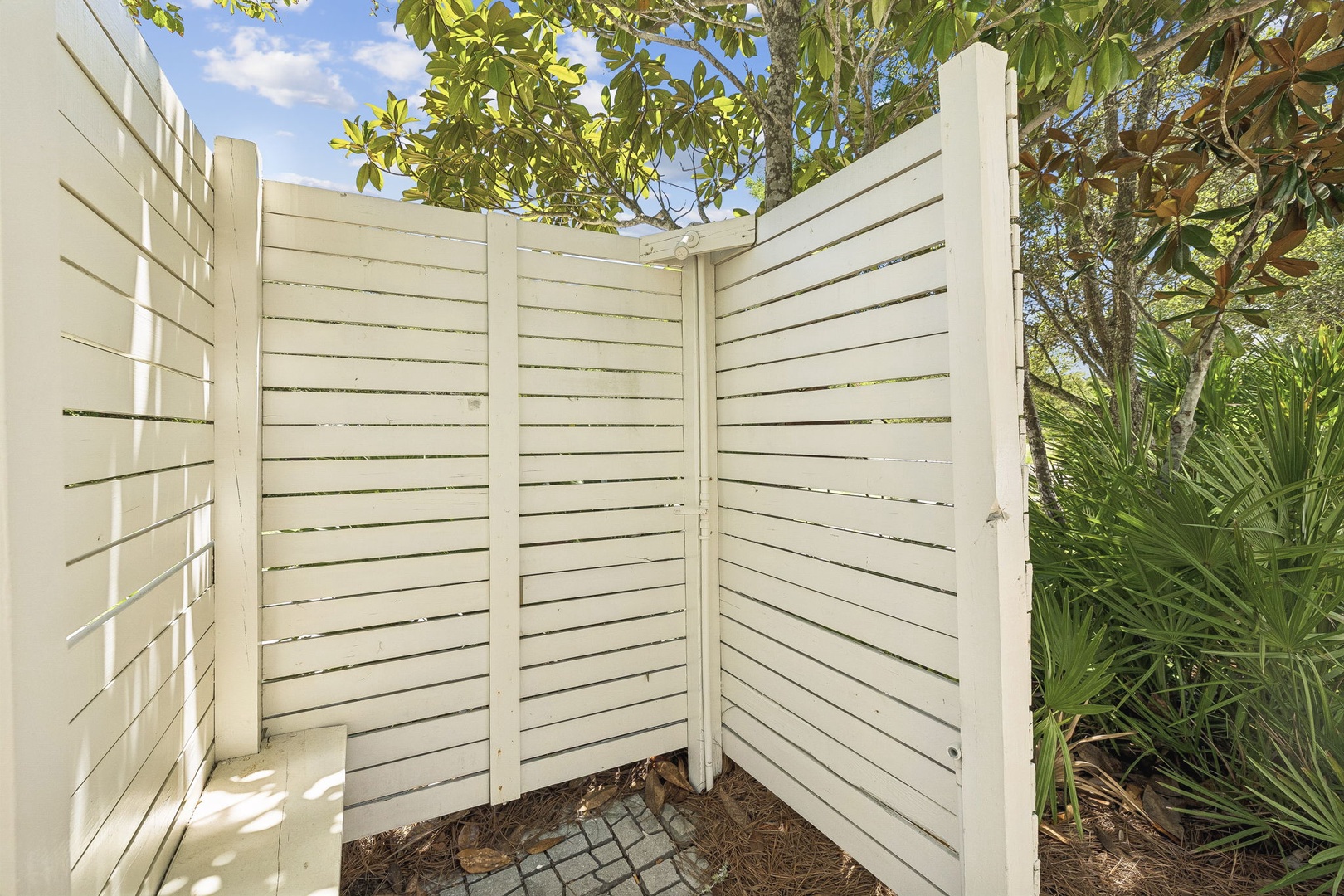 A white slatted outdoor shower framed by tropical palms and leafy greenery—perfect for rinsing off after time by the pool or a beach stroll.