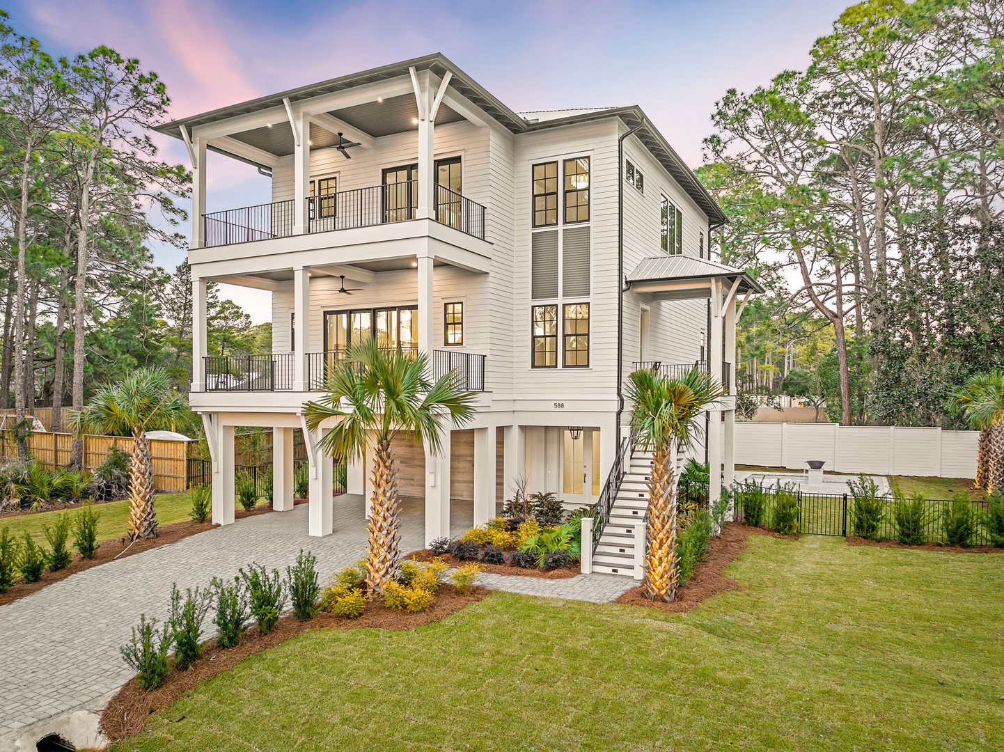 Modern three-story beach house with covered balconies and elevated design, surrounded by lush landscaping and mature trees in a peaceful coastal setting.