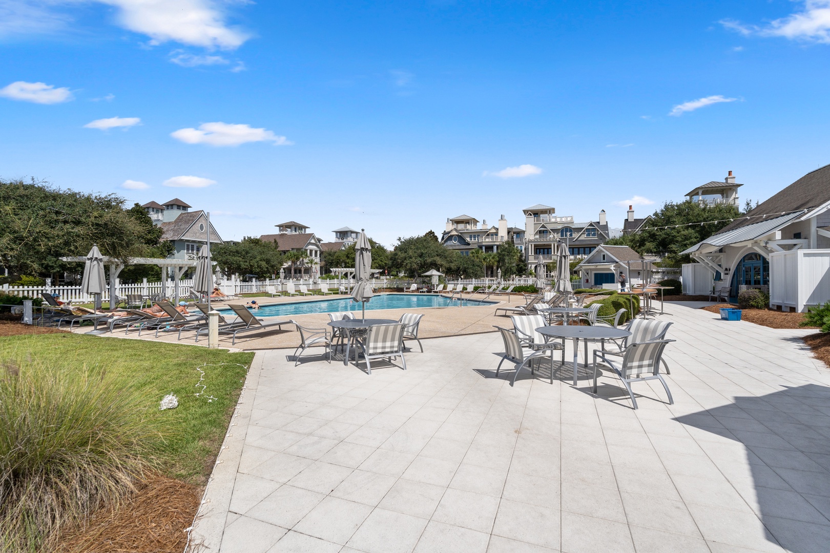 A sunlit pool area featuring lounge chairs, umbrellas, and outdoor dining tables set against a backdrop of charming coastal homes and lush greenery.