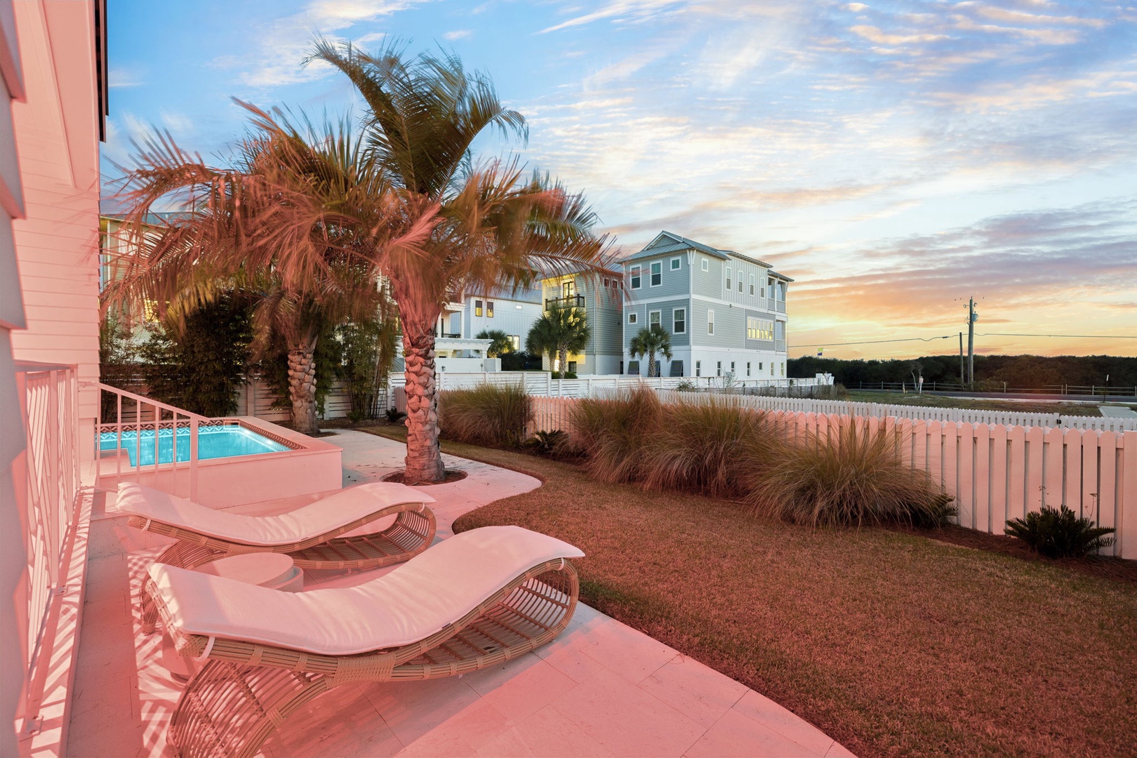 Coastal sunset paints the sky as palm trees frame the pool deck and neighboring beach houses in warm golden light.