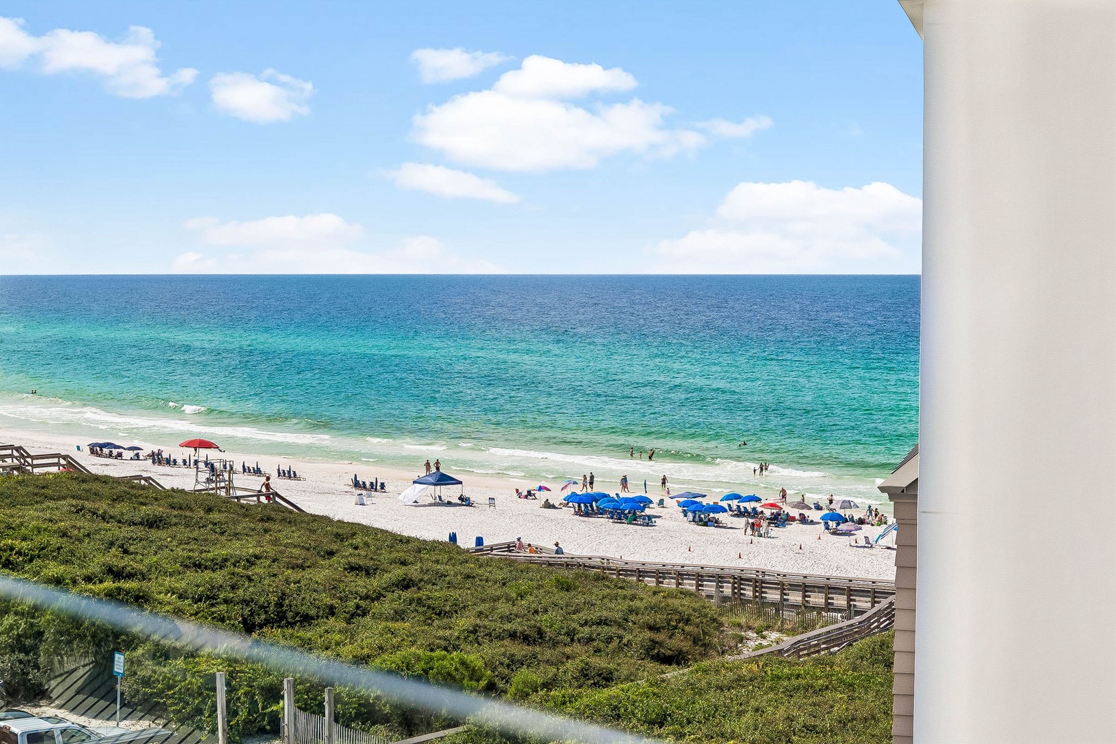 Elevated view captures a pristine stretch of white sand beach meeting turquoise Gulf waters, with beachgoers enjoying sunny skies and coastal dune vegetation framing the scene.