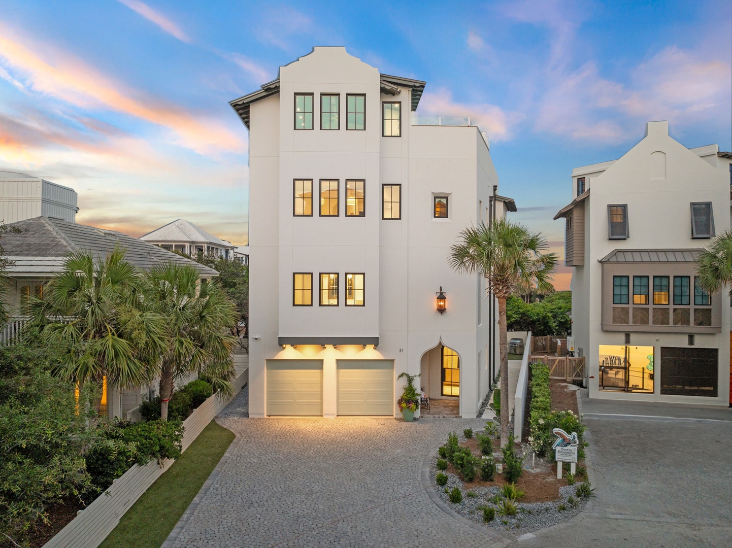 An elegant three-story coastal retreat nestled in a peaceful neighborhood, framed by palm trees and neighboring homes beneath a pastel sunset sky, welcoming you to a sophisticated beachside escape.