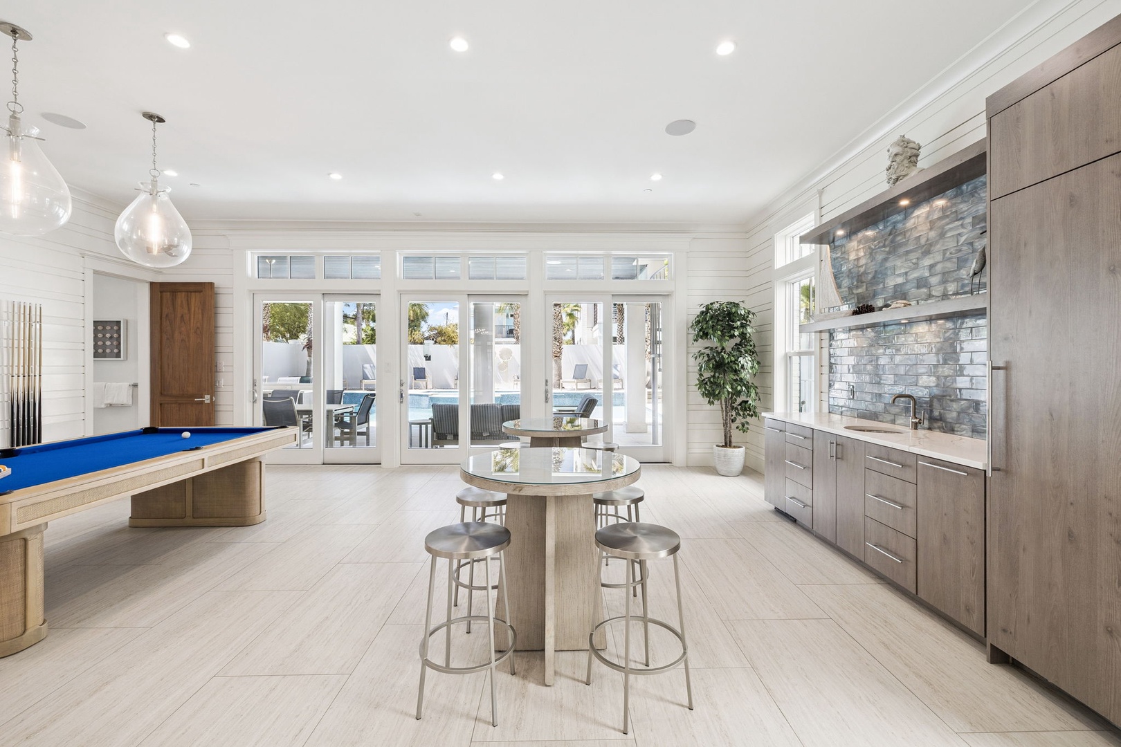 Step into your sophisticated entertainment space featuring a sleek kitchen, billiards table, and stunning pool views through floor-to-ceiling windows.