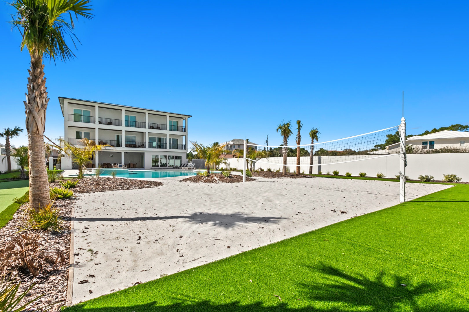 Modern three-story property with a spacious pool area, sandy volleyball court, and tropical palm landscaping under clear blue skies.