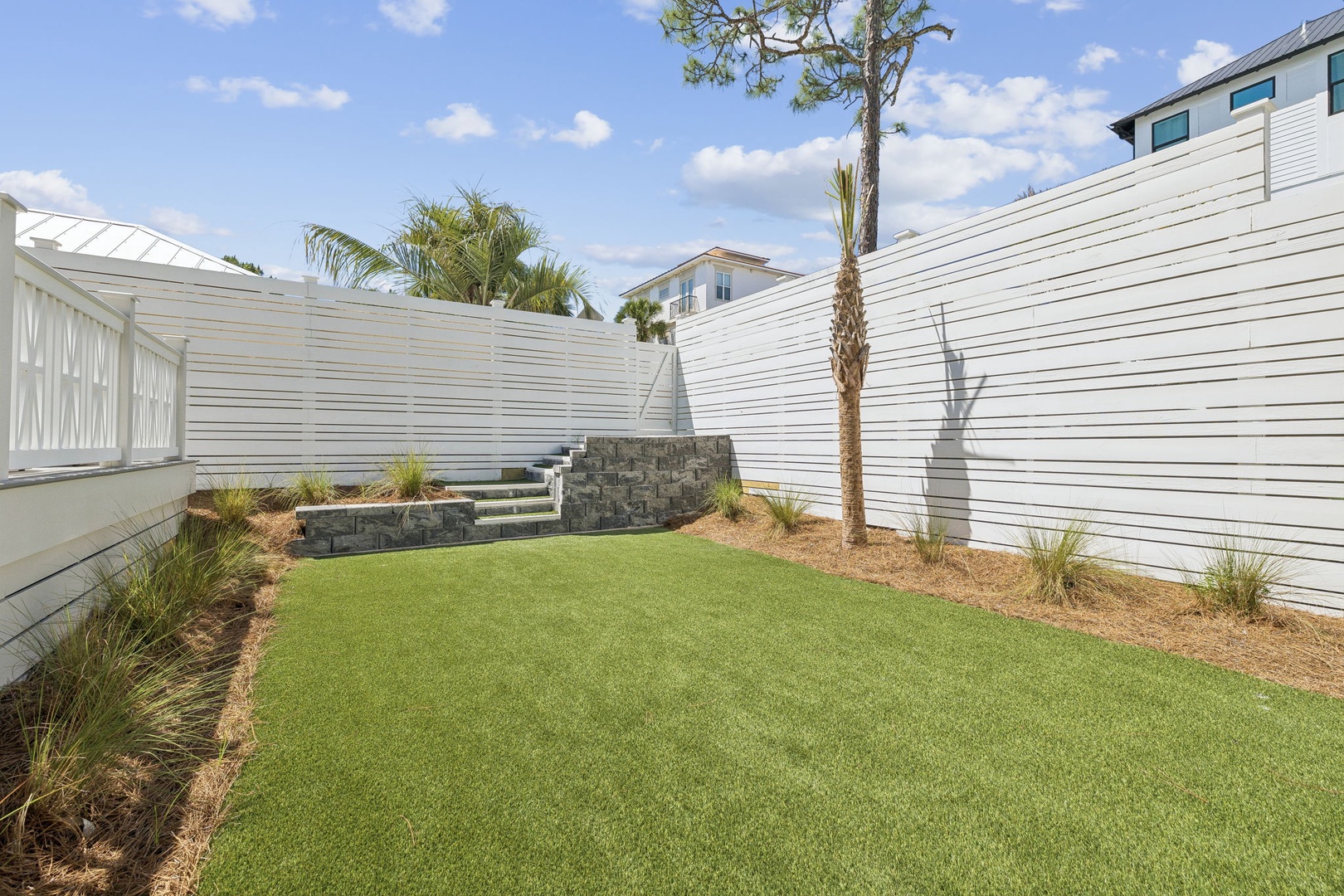 Your private backyard oasis features pristine artificial turf surrounded by tropical palms and modern stone accents, perfect for morning coffee or evening relaxation.