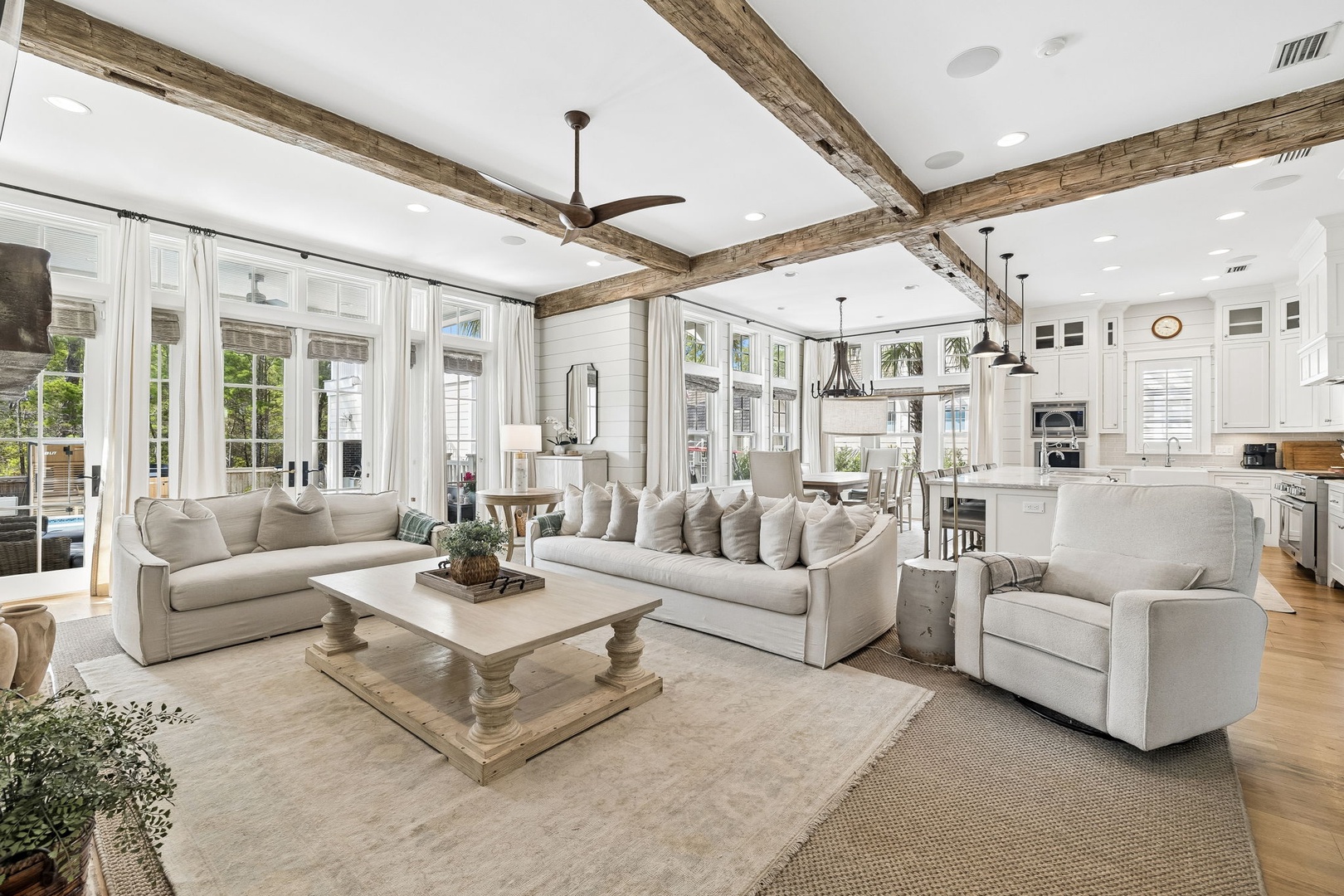 Sink into plush sofas beneath soaring beamed ceilings in this sun-drenched open-concept living area, where farmhouse charm meets modern elegance and expansive windows frame lush garden views.