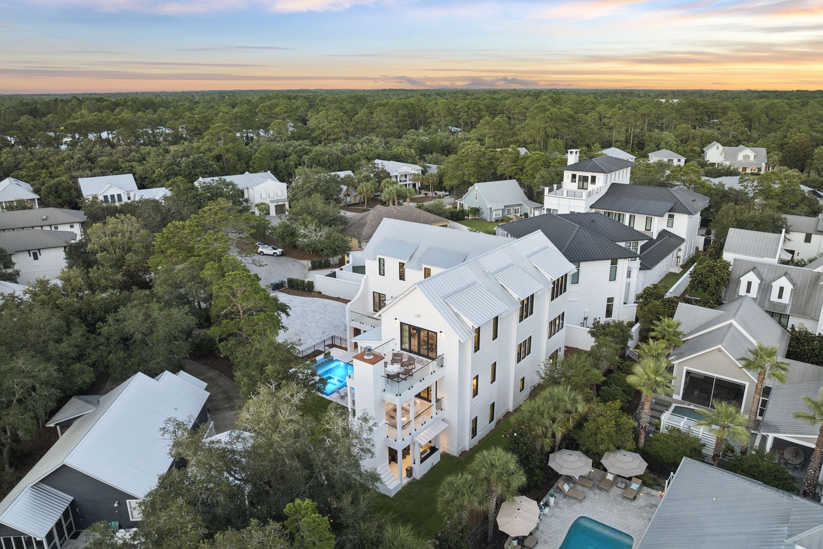 An elegant three-story coastal home nestled among lush trees in a serene residential neighborhood, captured at golden hour with the property's private pool and outdoor terraces visible.