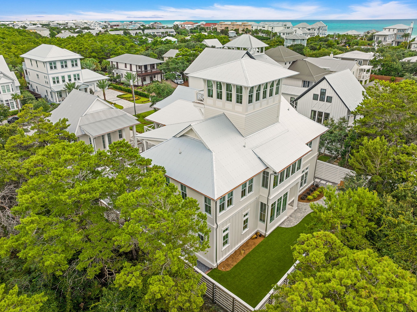 An aerial view captures this coastal property nestled among lush greenery, with the turquoise sea visible in the distance. The area offers a serene escape surrounded by natural beauty and neighboring homes.