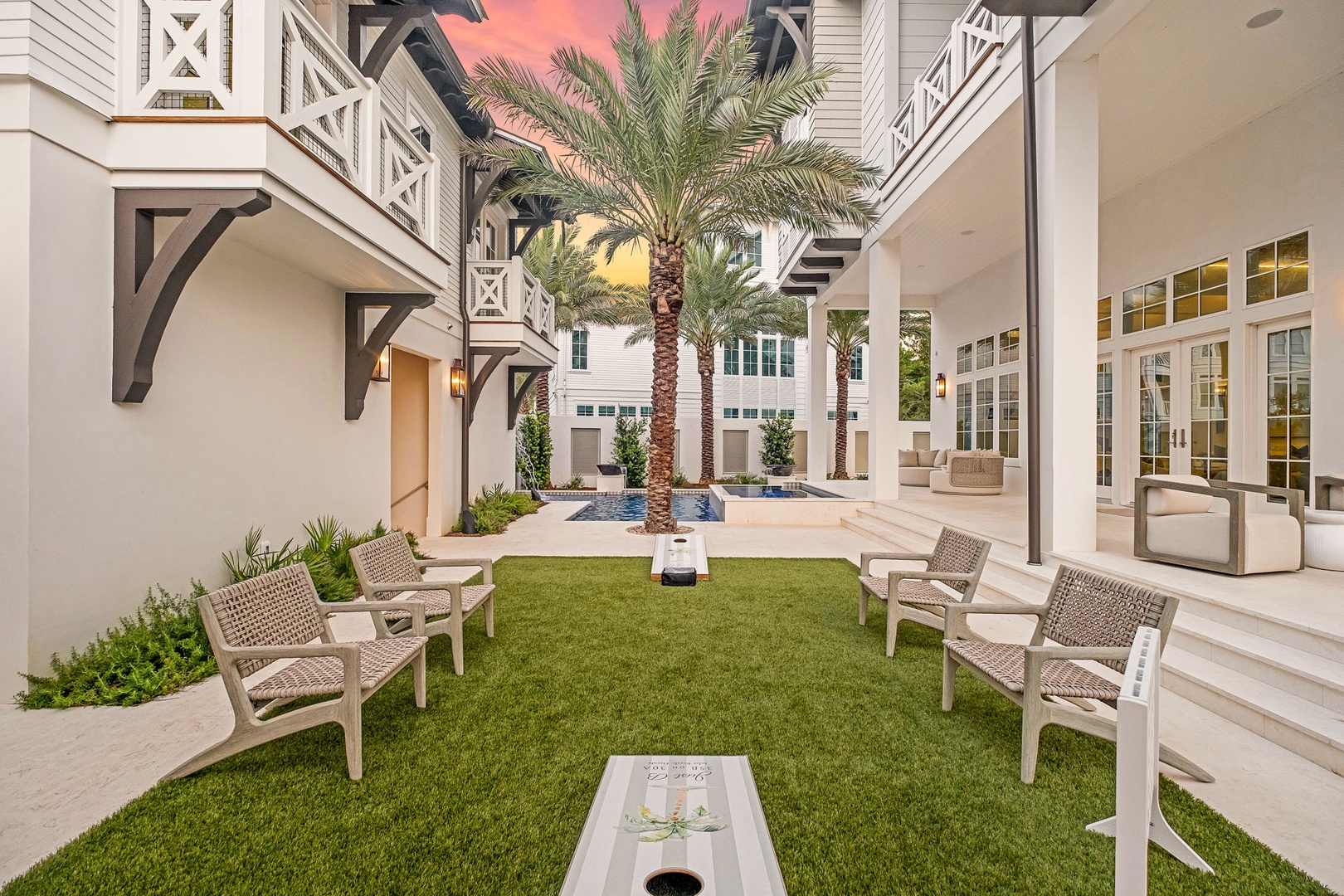 The property features an inviting inner courtyard with lush palm trees, green lawn, and comfortable seating areas framed by elegant white architecture and soft dusk skies.