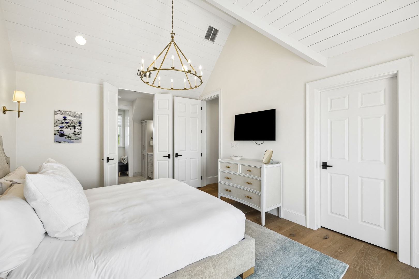 Picture yourself waking in this serene bedroom, where vaulted ceilings and soft white linens invite total relaxation. A statement chandelier adds charm, while the flat-screen TV and dresser offer comfort. Your private ensuite awaits beyond those doors.