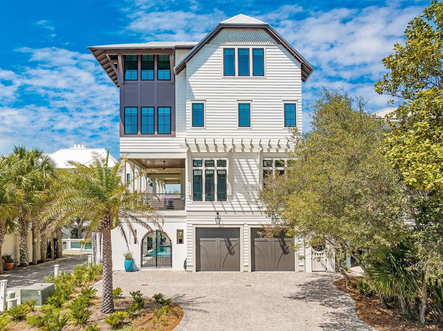 Three-story coastal vacation home with modern architecture, surrounded by palm trees and tropical landscaping under sunny skies.