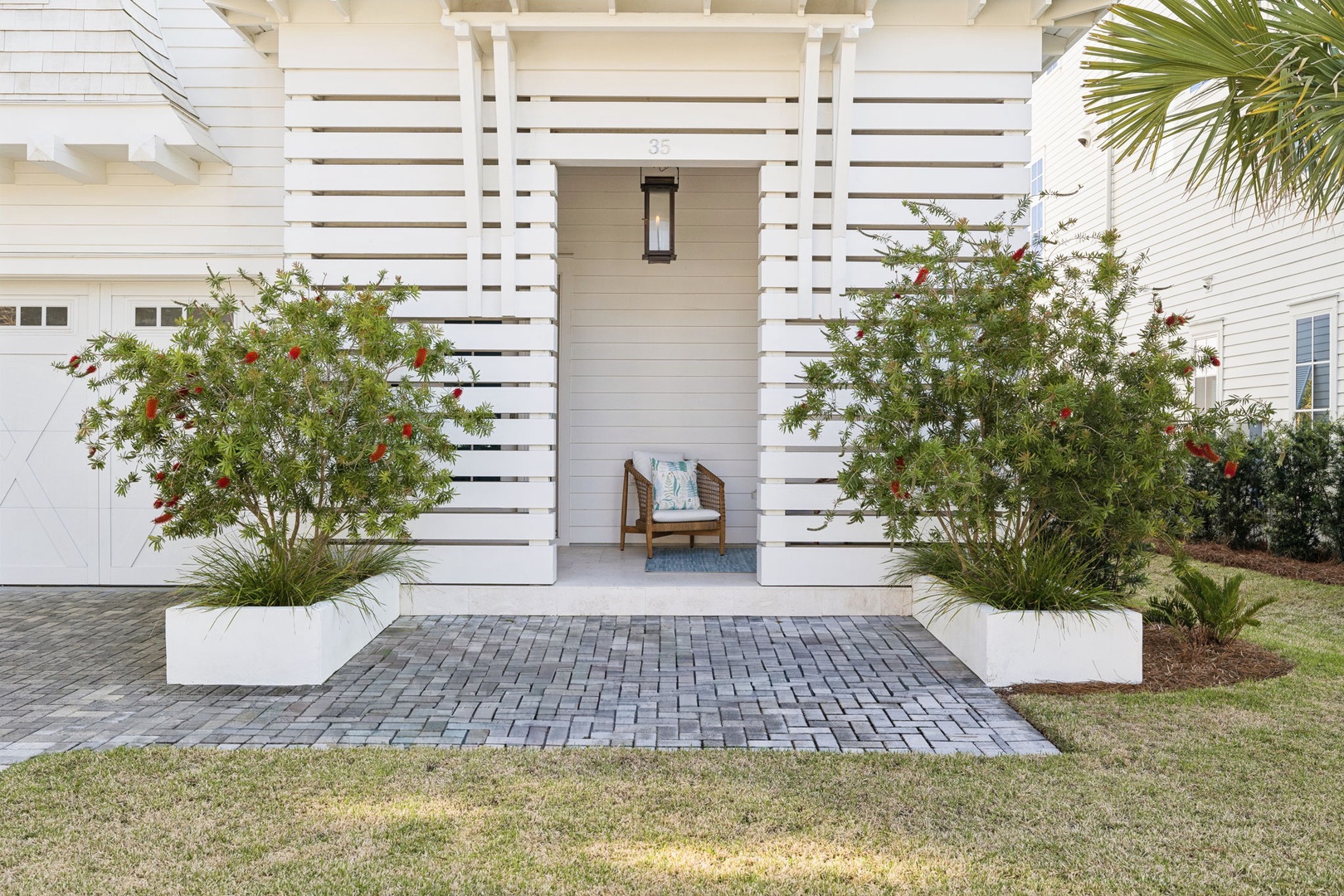 Modern coastal entrance showcases tropical landscaping with pomegranate trees and palm foliage surrounding a brick-paved courtyard.
