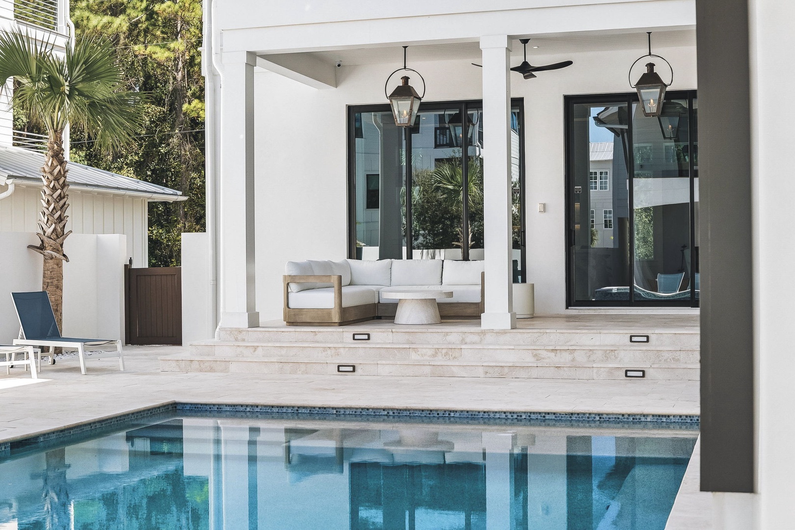 Chic coastal-modern outdoor pavilion overlooking a pristine swimming pool, offering seamless indoor-outdoor living where you can unwind on plush seating beneath elegant pendant lights, with lush palms framing your private retreat.