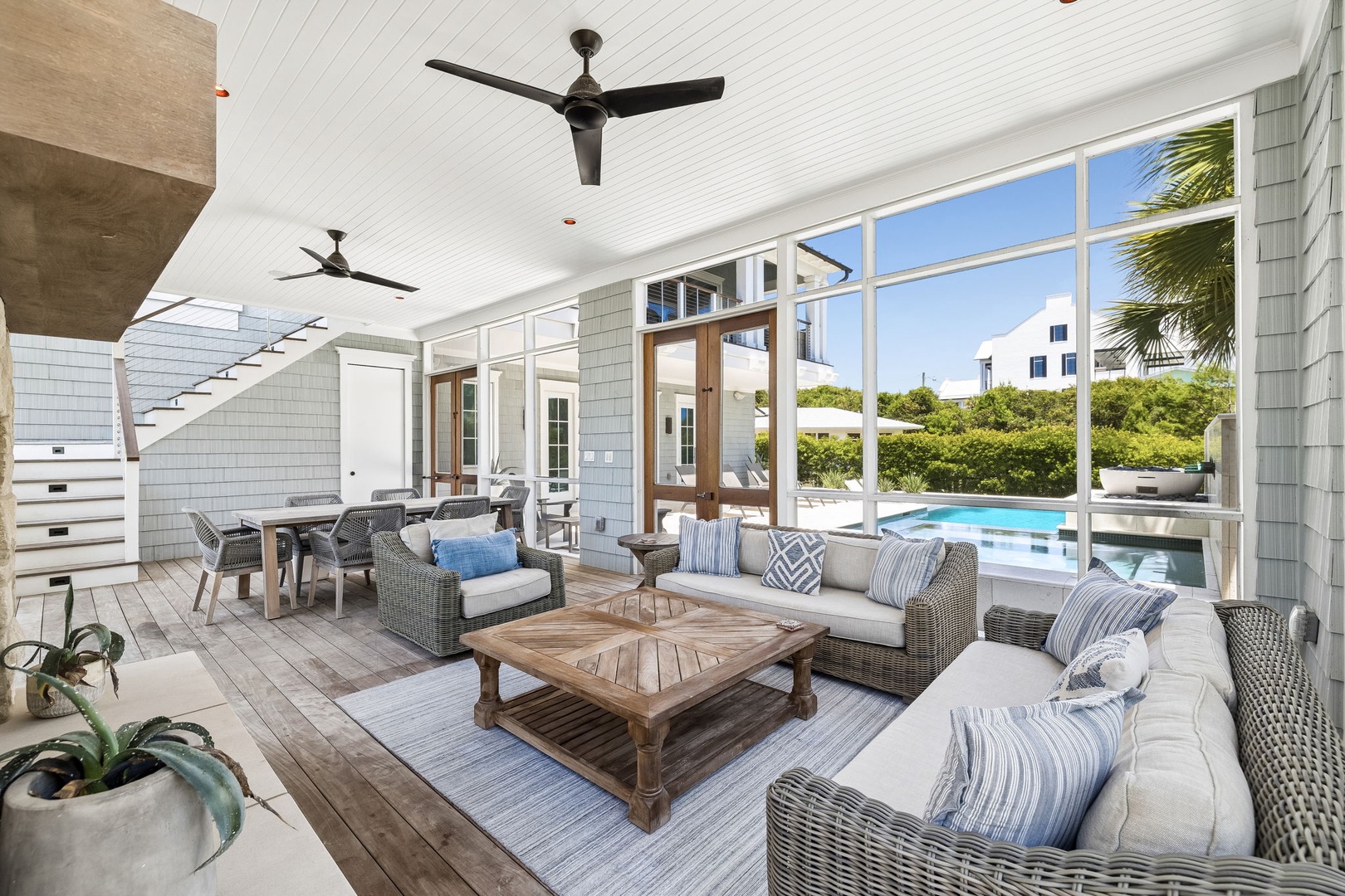 Covered outdoor living area features pool views, coastal-style lounge seating, and dining space, framed by white shiplap and ceiling fans for year-round comfort.