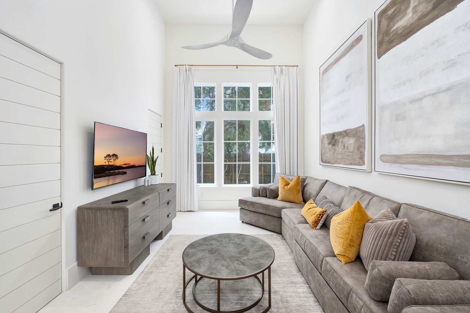 Sink into the plush gray sectional and unwind in your bright, airy living space, where soft pops of golden yellow and a gentle breeze from the ceiling fan invite total relaxation.