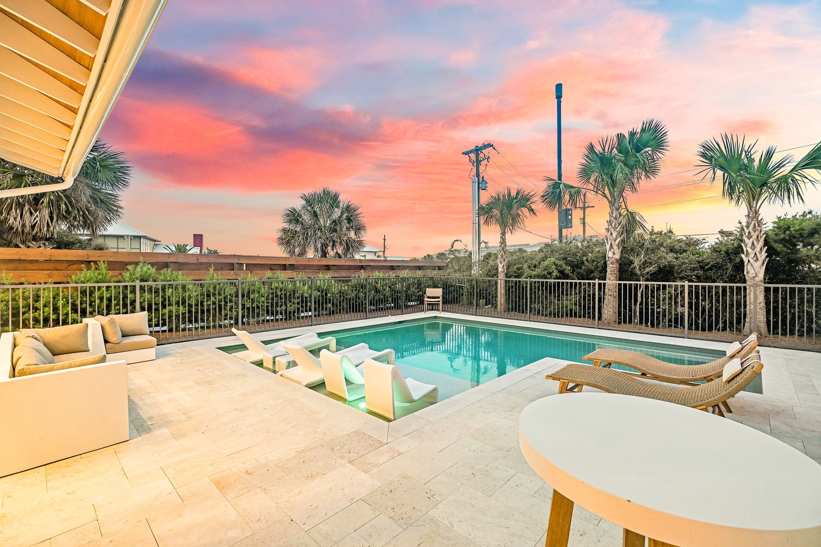 A tropical oasis featuring a sparkling pool, palm trees, and stunning sunset views painting the sky in coral and lavender hues above the surrounding area.