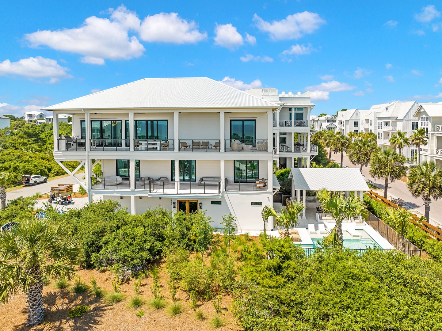 Modern coastal vacation home featuring multiple covered balconies and wraparound porches, nestled among tropical palms and lush landscaping in a sunny beachside neighborhood.
