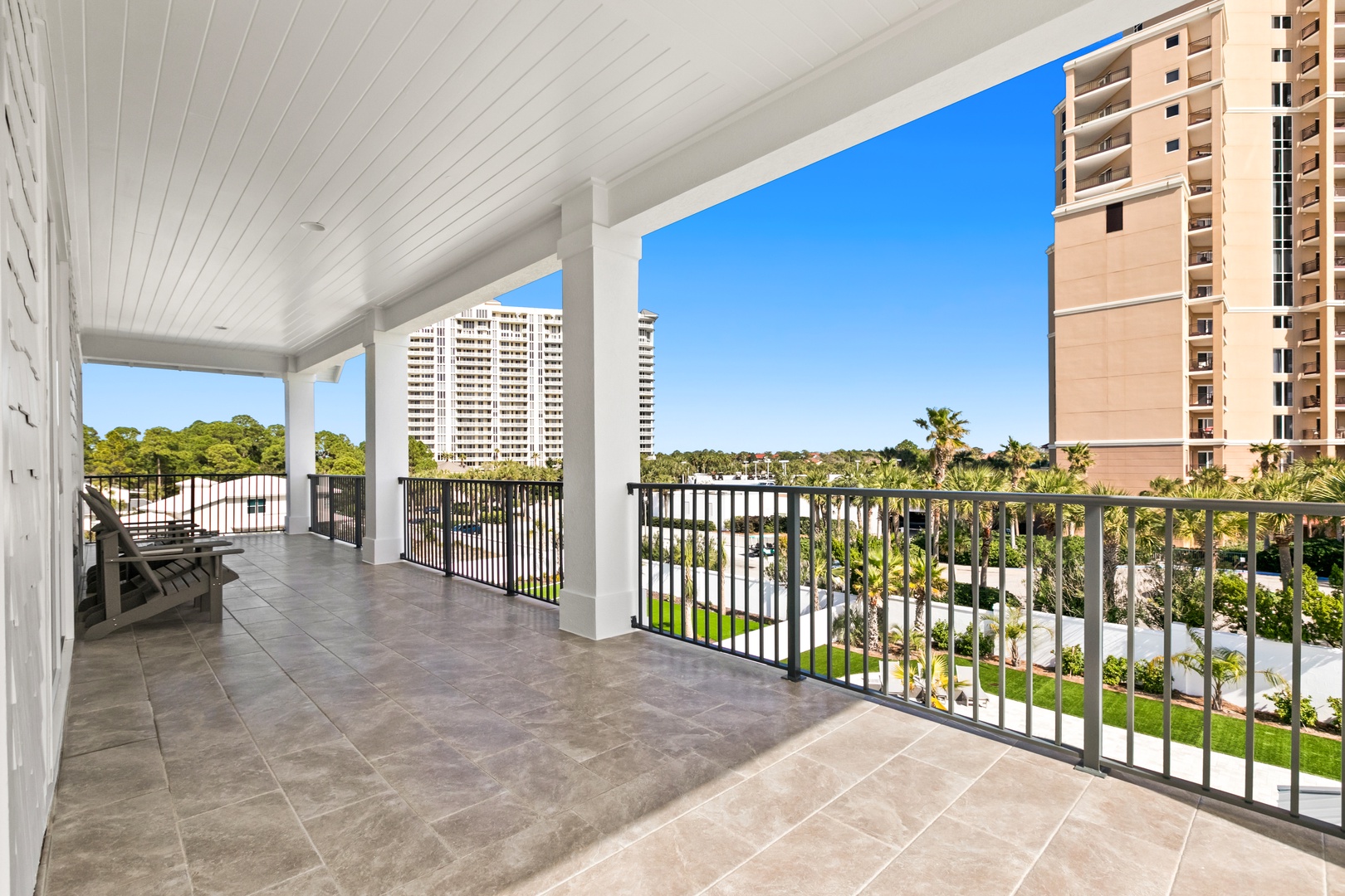 A spacious covered balcony overlooks a peaceful waterfront and lush landscaped grounds, framed by blue skies and swaying palm trees—your private outdoor retreat for morning coffee or evening sunsets.