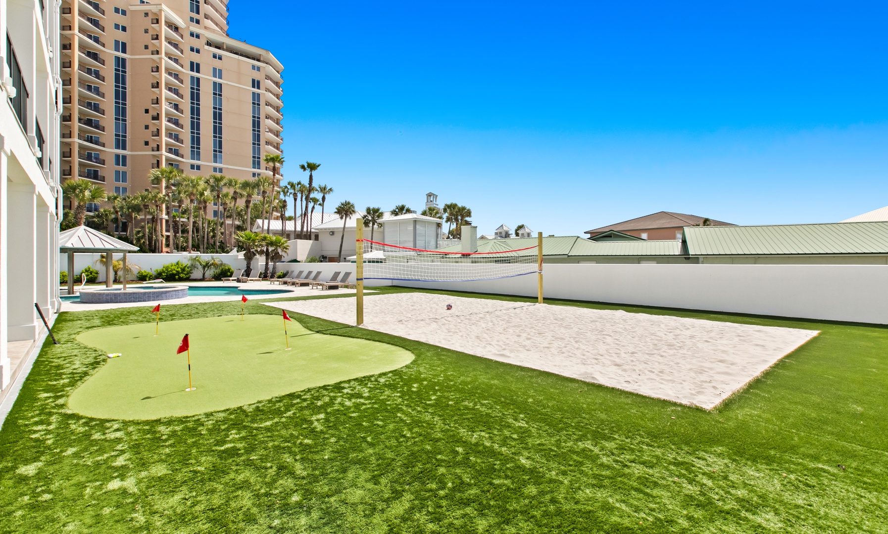 Recreational area with a small putting green, sand volleyball court, and palm trees in a coastal resort setting.
