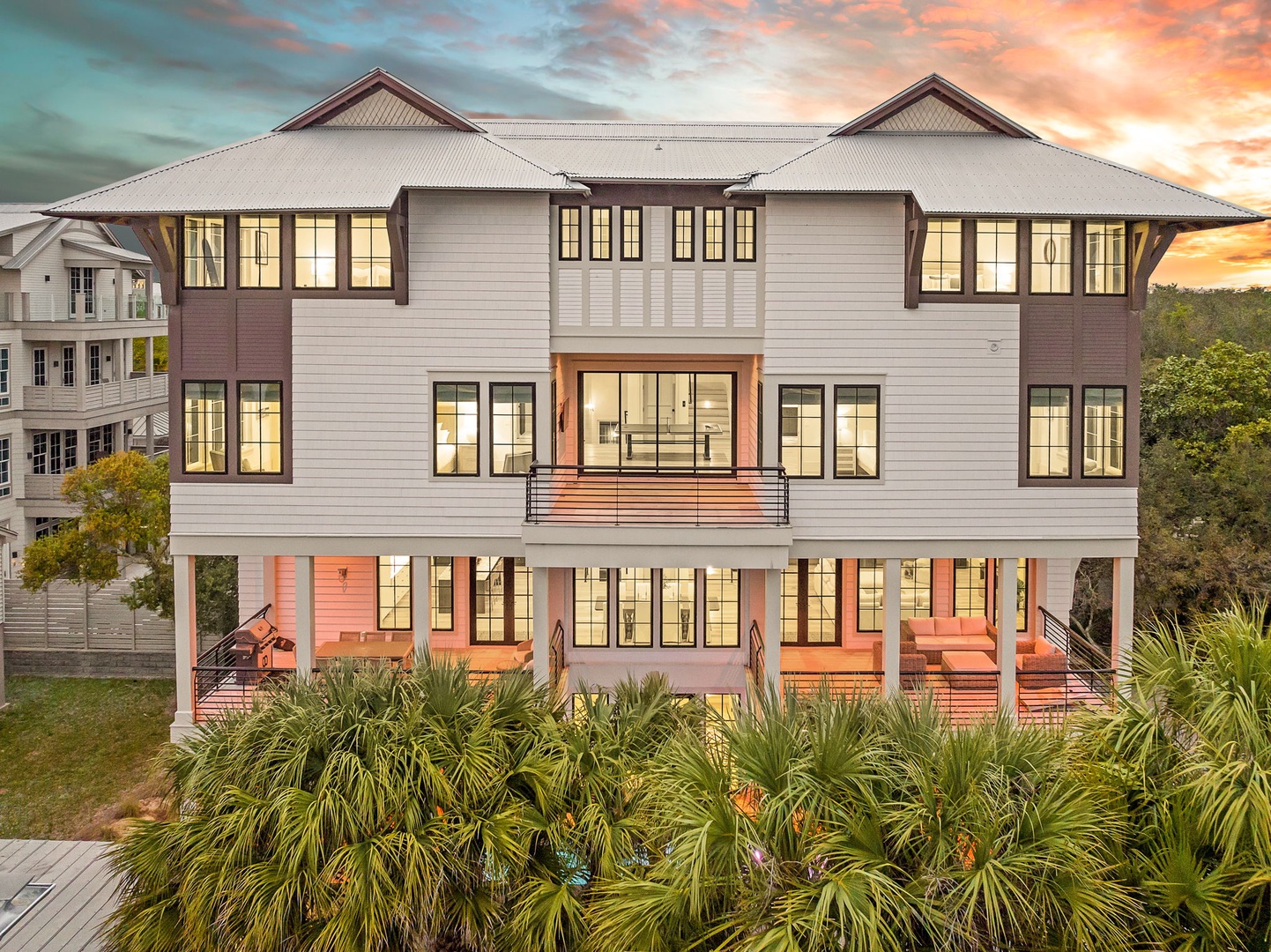 Stunning coastal property featuring modern architecture with warm lighting against a breathtaking sunset sky, surrounded by lush tropical landscaping.