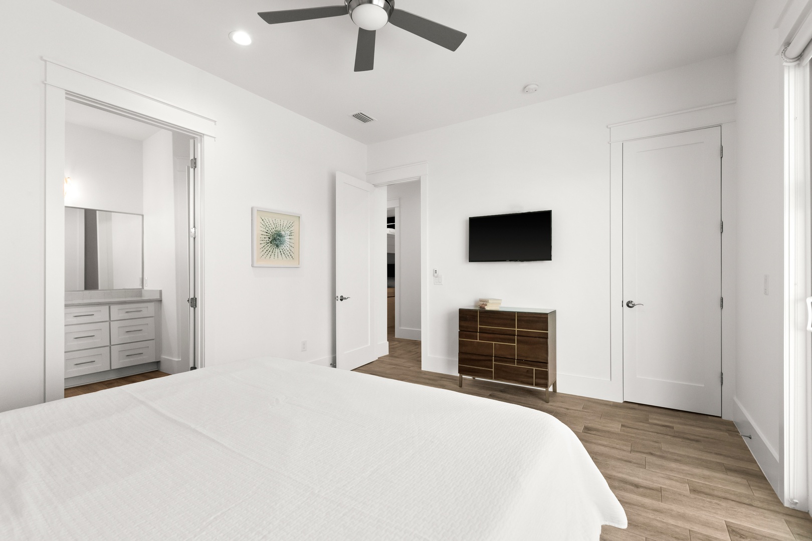 Step into your serene, sun-filled bedroom with crisp white linens, warm wood flooring, and a ceiling fan to keep you cool. The mounted TV and modern dresser offer comfort and convenience for your stay.