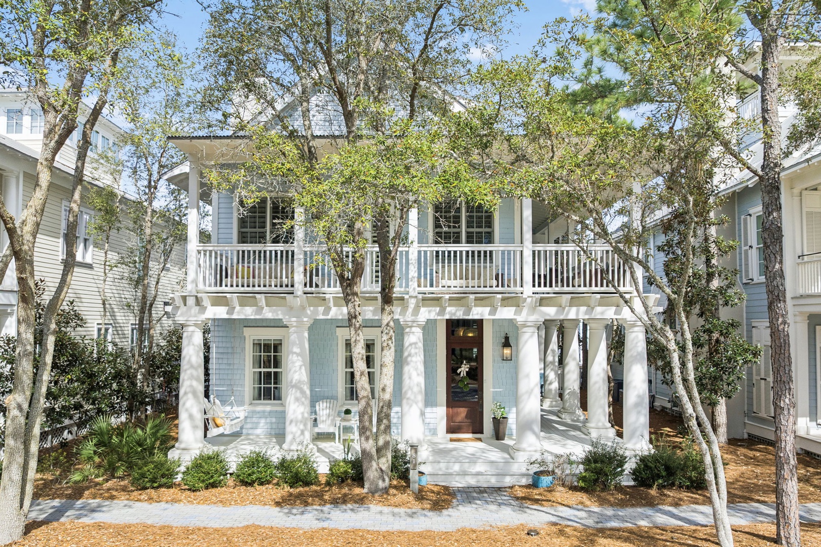 Charming coastal-style cottage with white columns and wraparound porches nestled among mature trees in a peaceful neighborhood setting.
