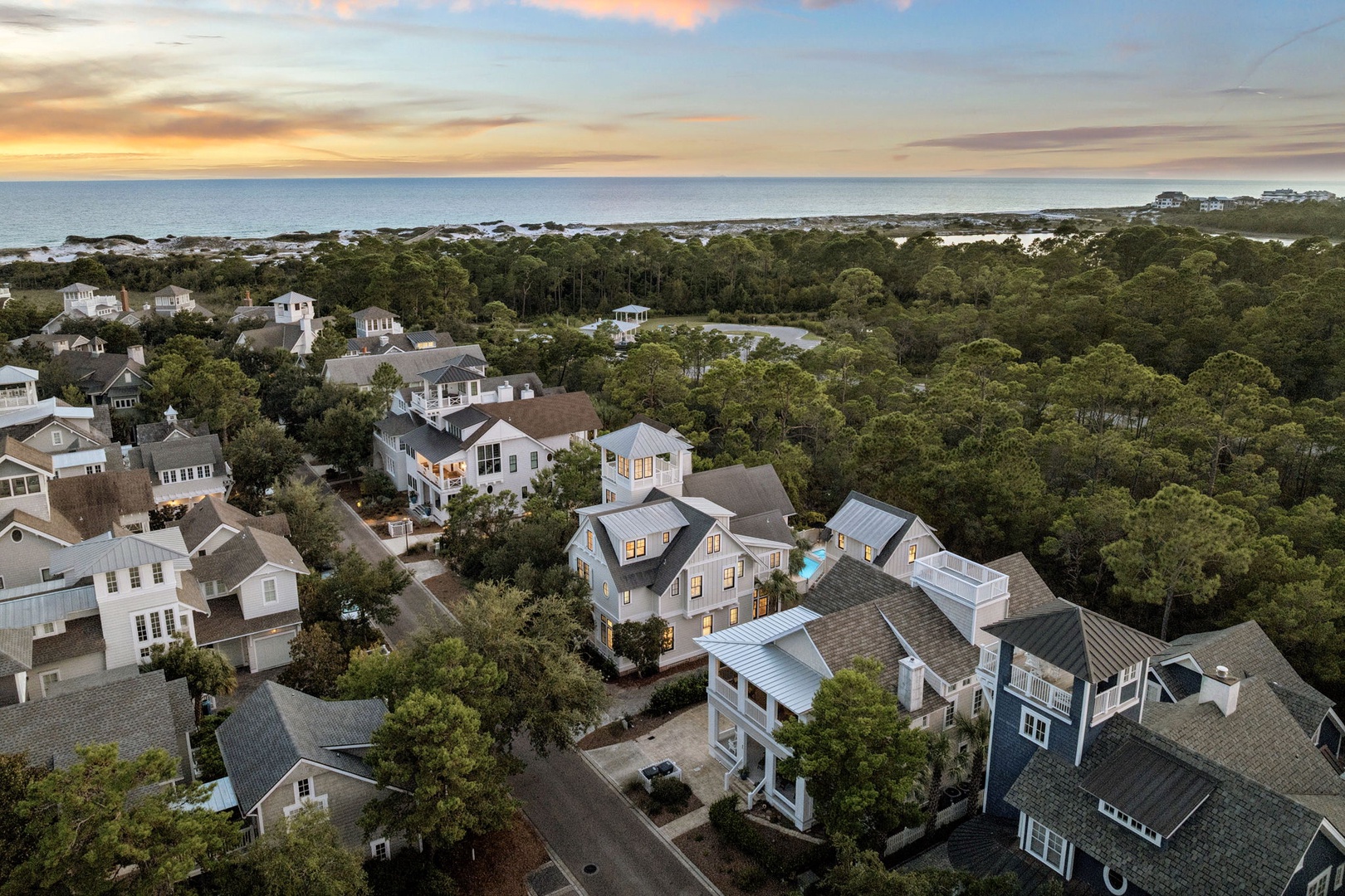 An aerial view showcases the coastal community nestled among lush trees, with the ocean stretching to the horizon and a stunning sunset painting the sky in soft pastels.