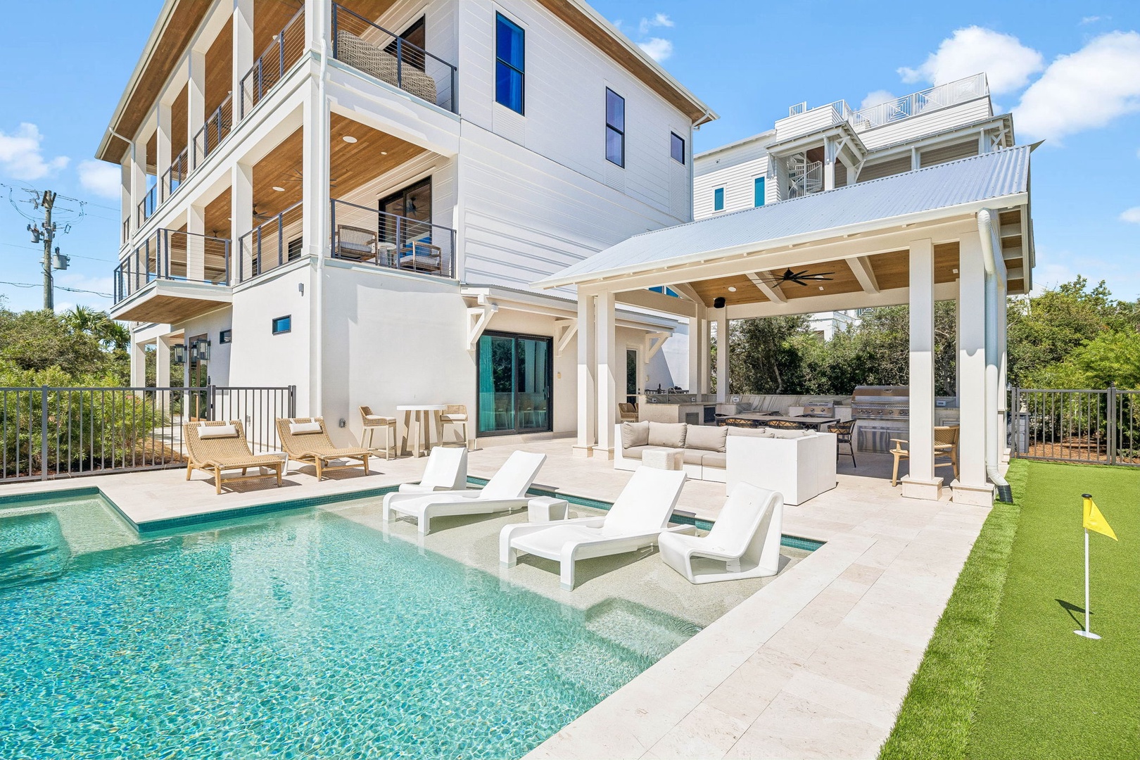 A stunning three-story property featuring pristine white architecture, a sparkling private pool with sun loungers, covered outdoor lounging areas, and a private putting green—everything you need for a luxurious coastal getaway.