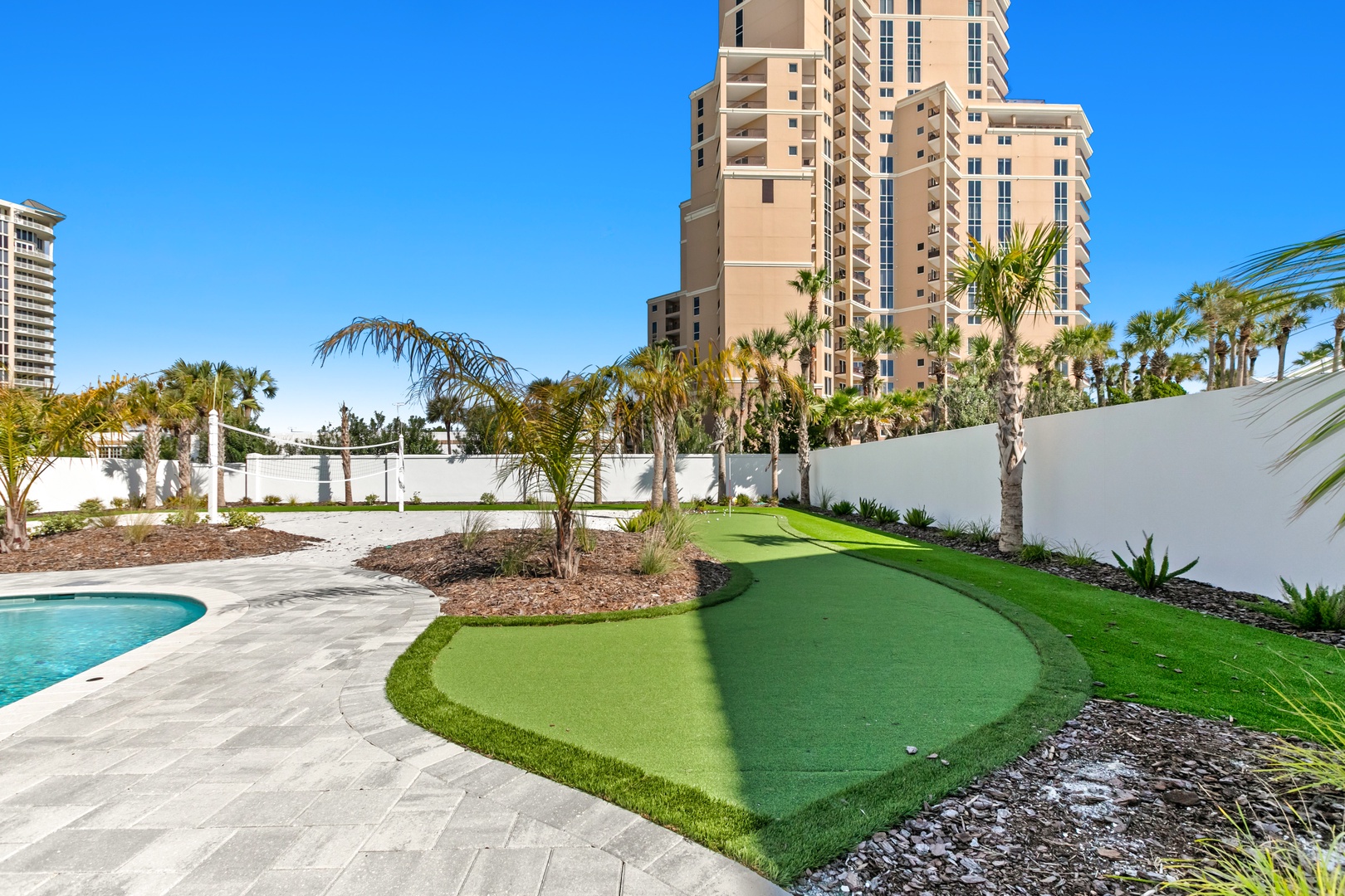 The property features a miniature golf course with putting green, palm trees, and pool area, set against a backdrop of nearby high-rise buildings in a coastal resort setting.