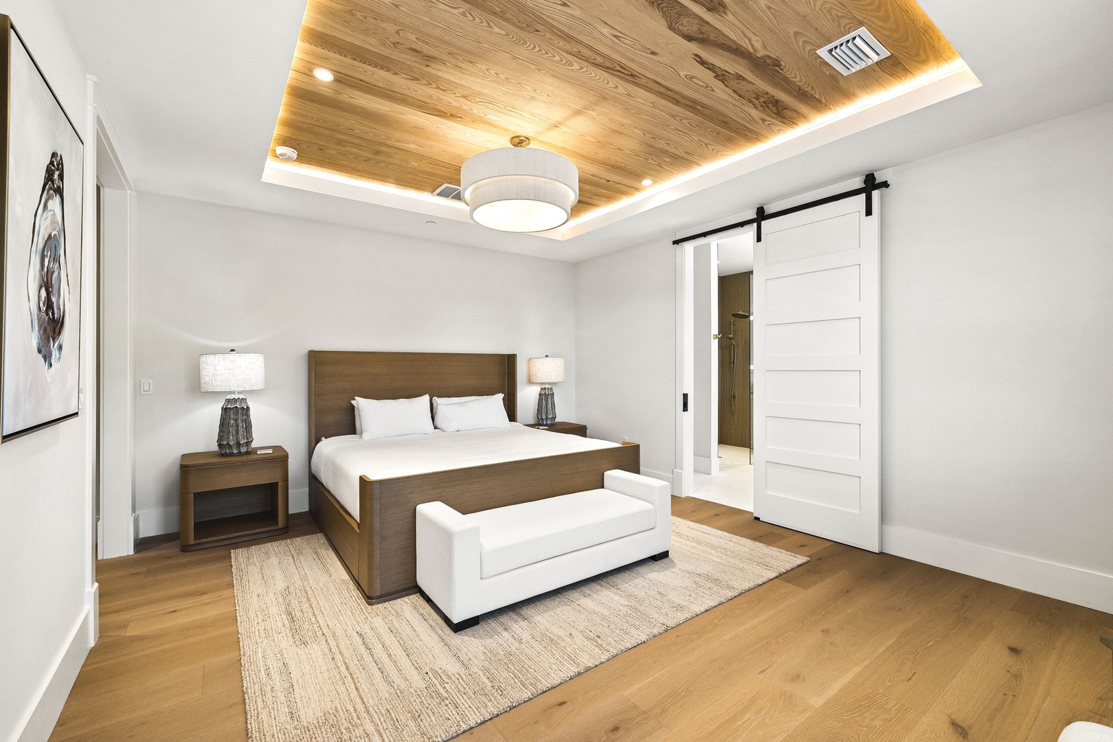 Drift off beneath a stunning wood-paneled ceiling lit by warm ambient light. Your plush king bed, flanked by elegant nightstands, invites pure relaxation. A cozy bench and sliding barn door add charm to your restful retreat.