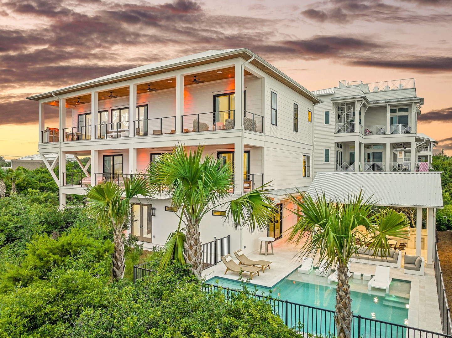 Modern three-story coastal retreat featuring palm trees, a sparkling pool, and multiple balconies set against a stunning sunset backdrop.