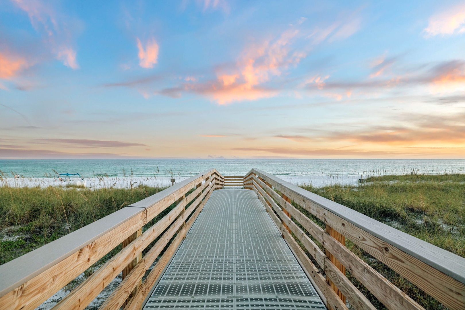 A wooden boardwalk leads through coastal dunes toward the calm ocean, with soft evening light painting the sky in golden and pastel tones.