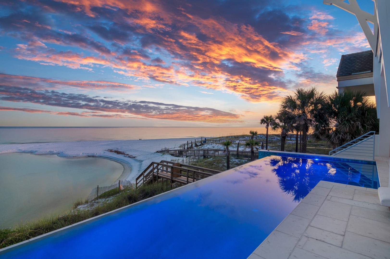 A stunning coastal property featuring an infinity pool overlooking peaceful waters, framed by swaying palms and dramatic sunset skies painting the horizon in brilliant coral and gold.