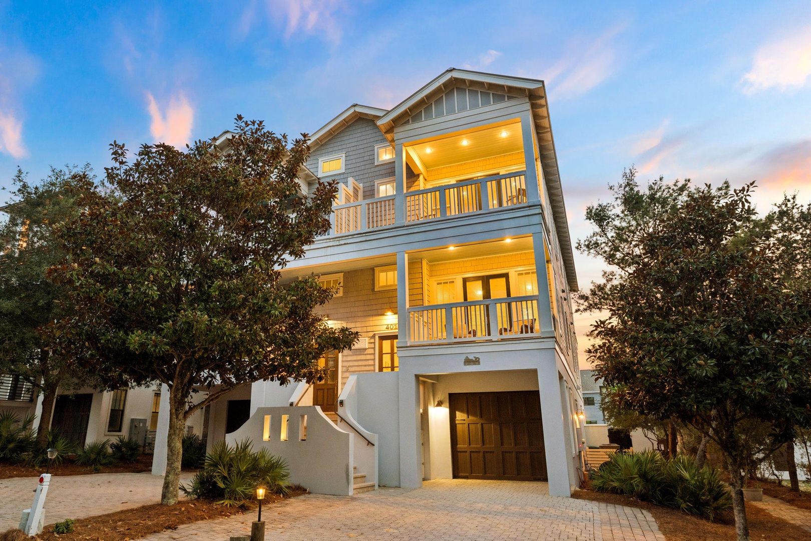 A stunning three-story coastal home welcomes you with warm evening light spilling from spacious balconies, framed by lush greenery and a charming paver driveway—your perfect beach retreat awaits.