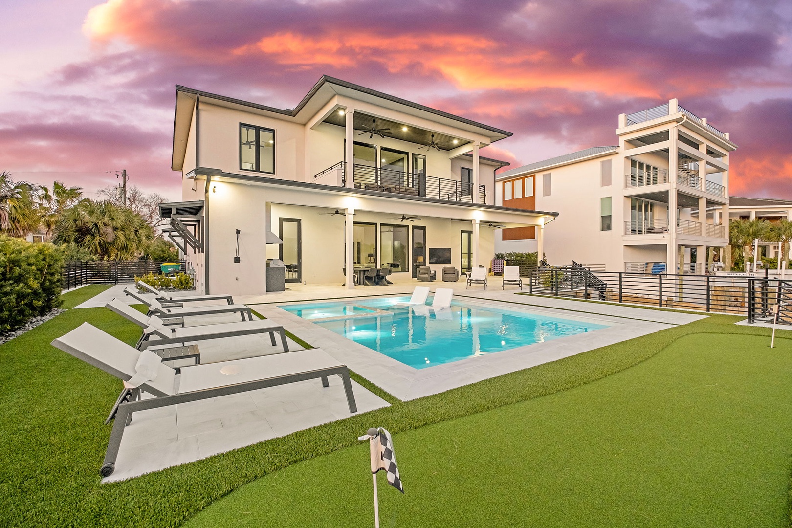 Modern beachside villa with sparkling pool and manicured lawn under dramatic sunset skies, showcasing contemporary architecture in coastal residential area.