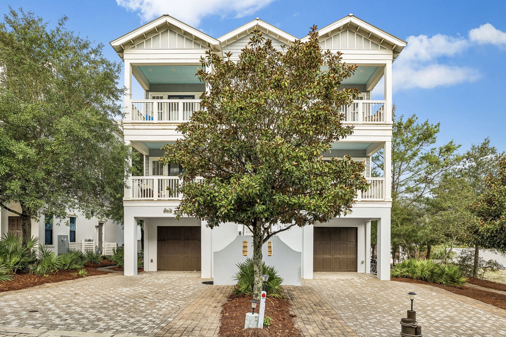 This elegant coastal duplex features white architectural details, spacious covered balconies, and mature landscaping framed by lush greenery and blue skies.