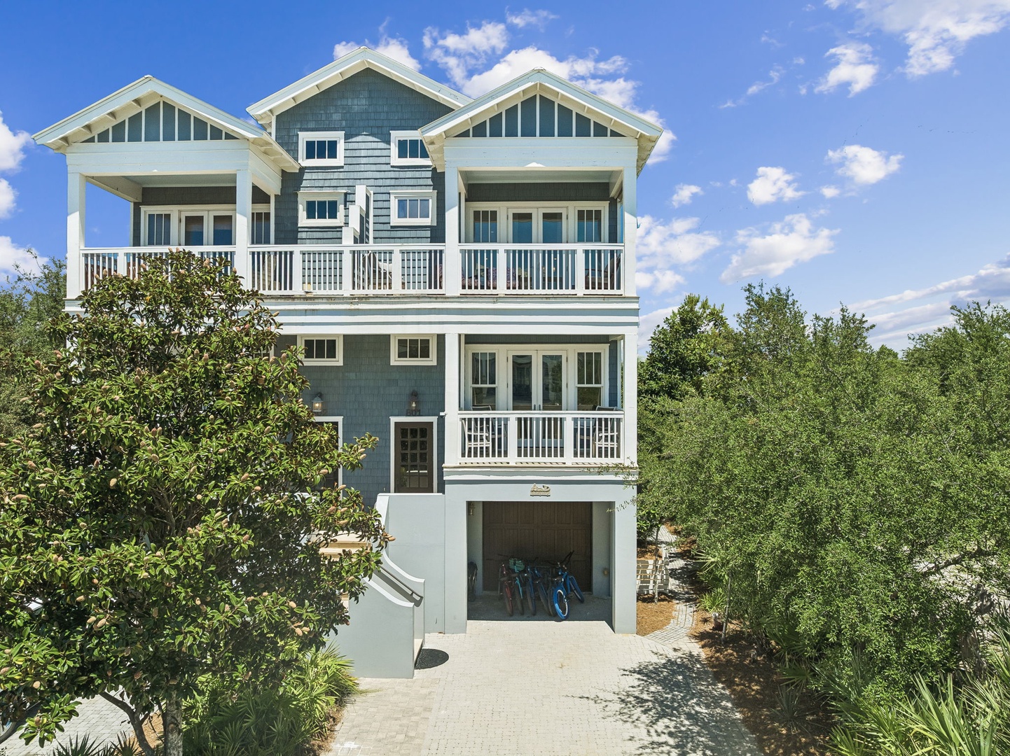 A charming coastal-style beach house featuring multiple balconies and open-air porches, nestled among lush greenery with convenient ground-level parking and bike storage.