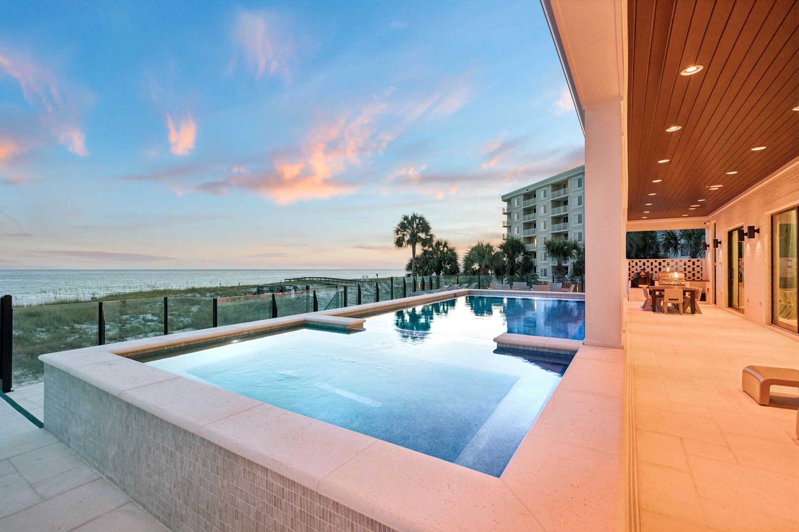 A stunning beachfront property features an infinity pool overlooking the ocean at dusk, complemented by a covered outdoor dining area and surrounded by swaying palms and coastal beauty.