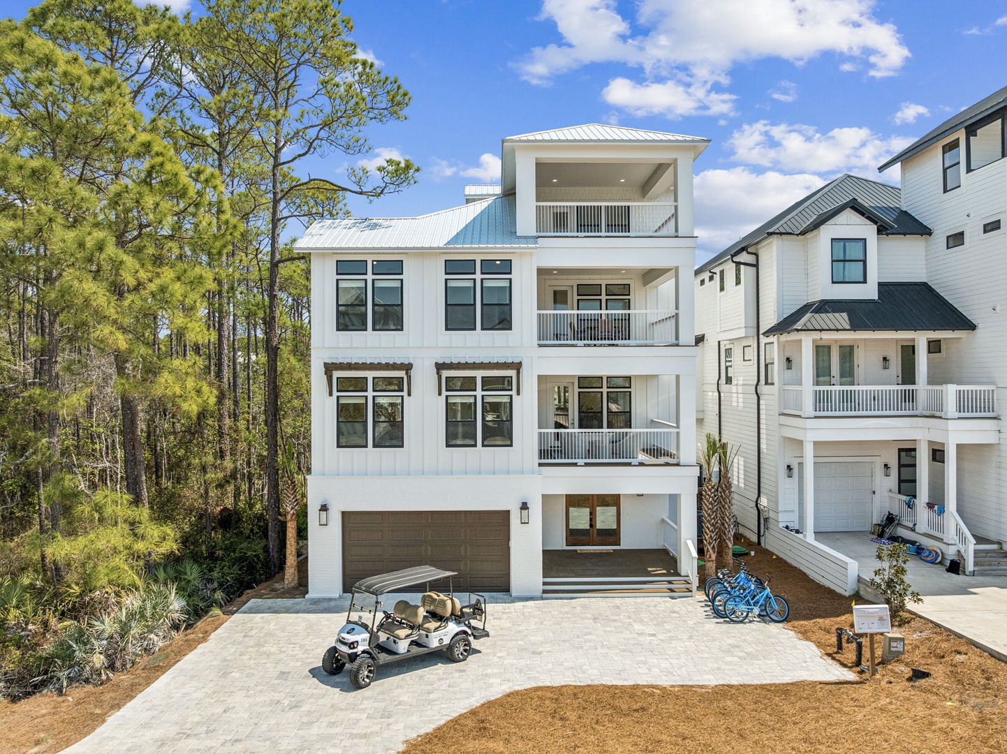 Modern three-story coastal home with multiple balconies and covered parking, surrounded by pine trees in a peaceful residential neighborhood.