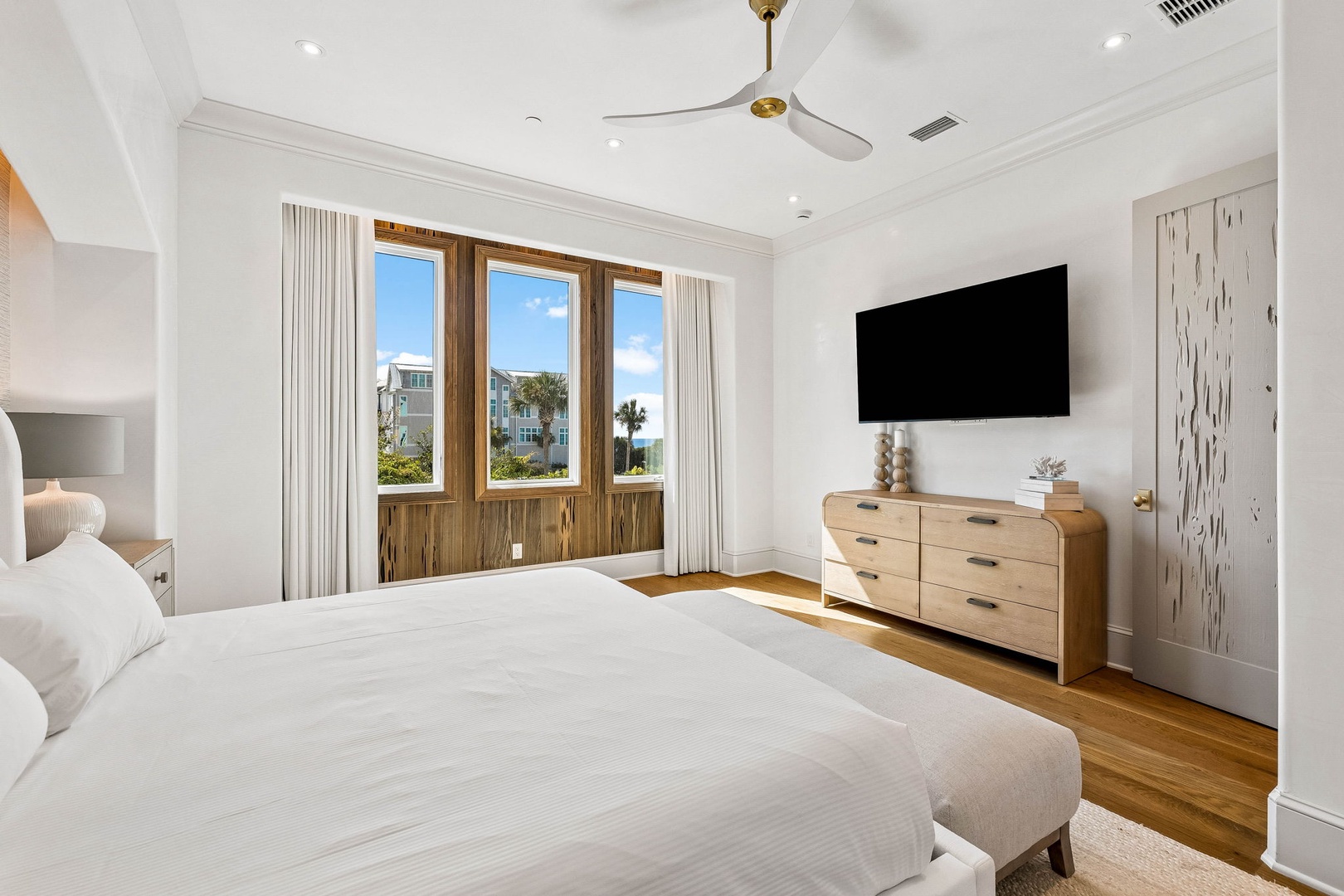 Wake up to palm trees and coastal views through triple wood-framed windows. Drift off in this airy, sunlit bedroom with crisp linens, a ceiling fan, and modern entertainment at your fingertips.