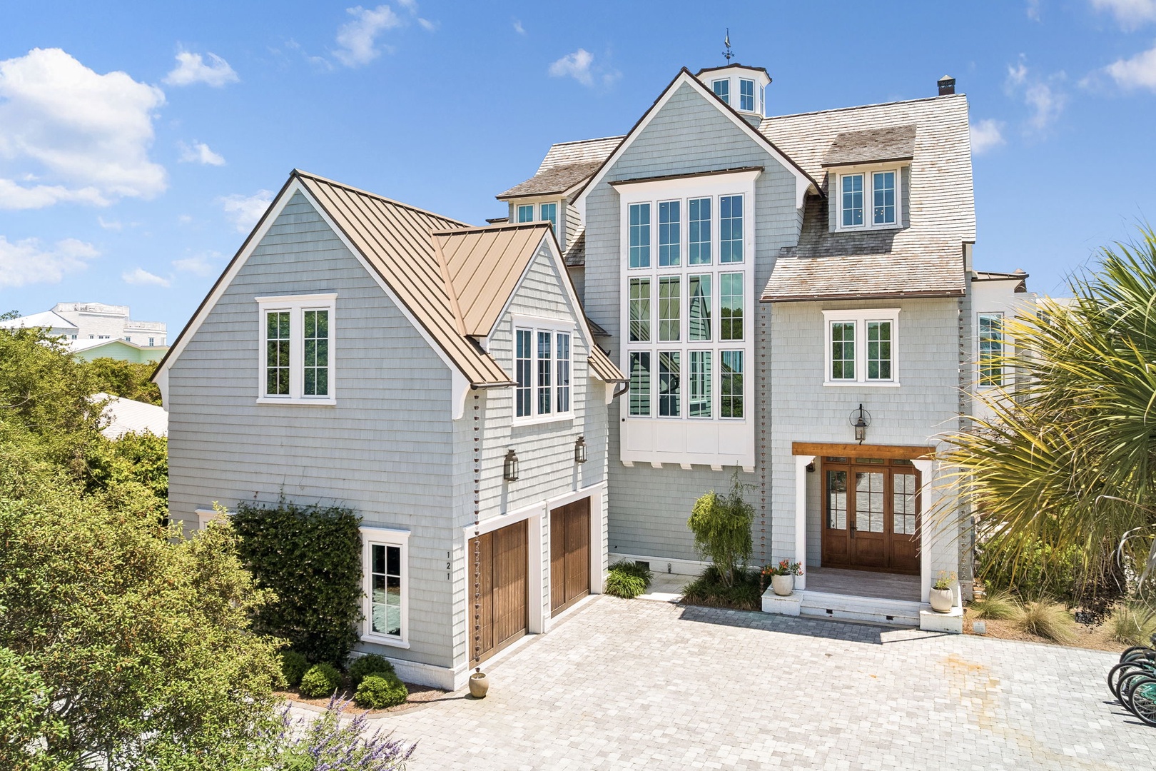 A beautiful coastal-style vacation home featuring light gray siding, elegant copper roofing, and a welcoming entrance surrounded by lush landscaping.
