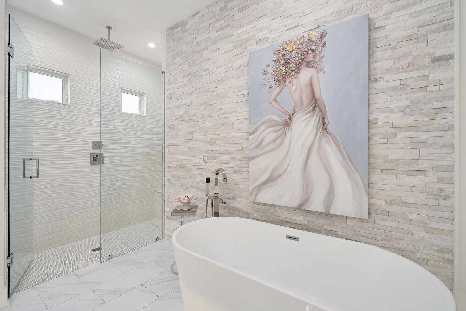 Step into a spa-like sanctuary featuring a freestanding soaking tub, sleek walk-in shower, and elegant stone accent wall with stunning artwork.
