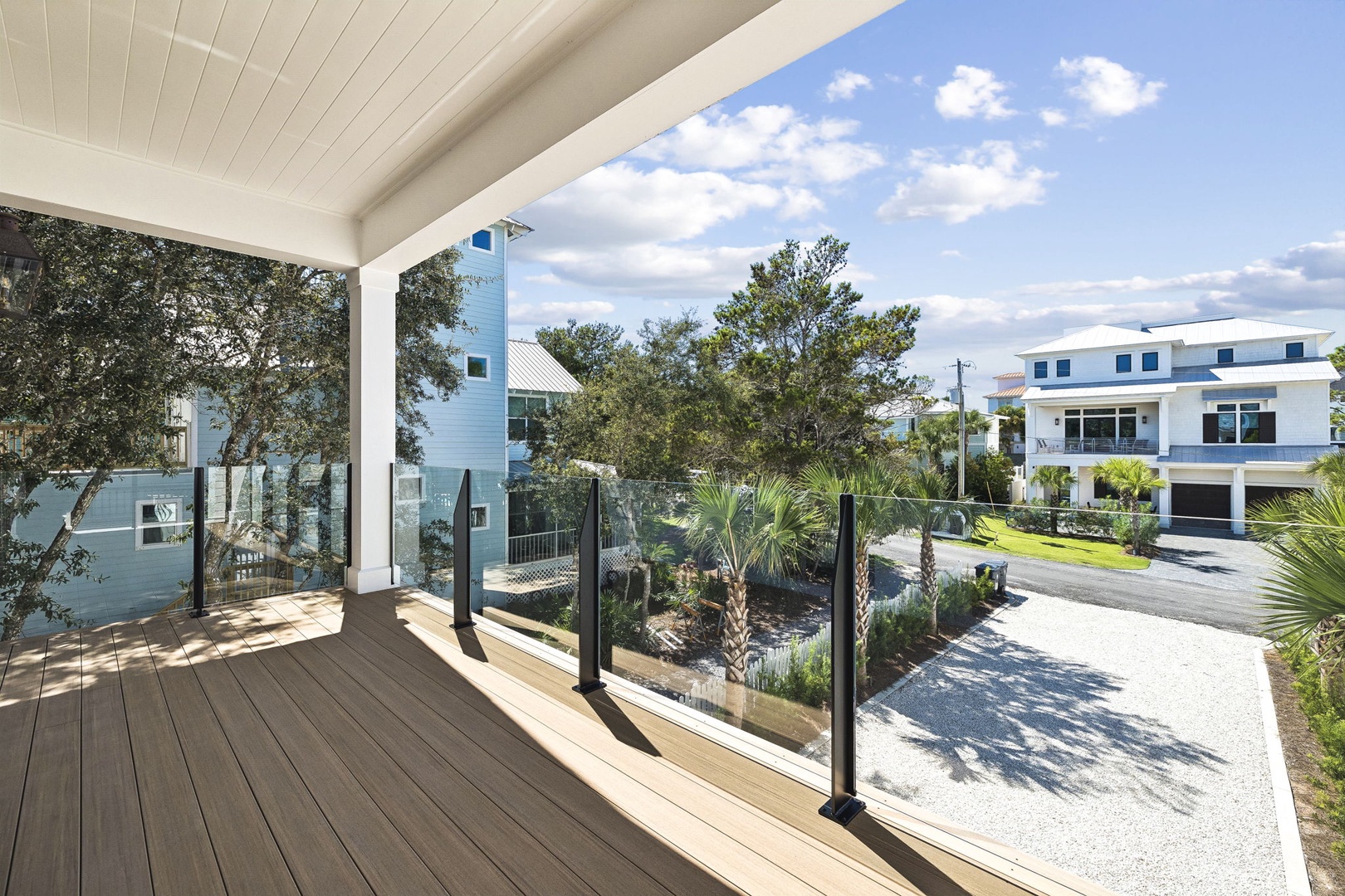 A covered balcony with modern glass railings overlooking a peaceful coastal neighborhood, surrounded by palm trees and neighboring beach homes under a bright blue sky.